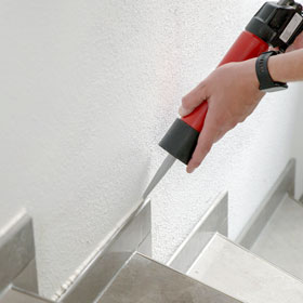 Hände mit Silikonspritze, die gerade eine Acrylfuge zwischen Treppensockel und Wand ausspritzt
